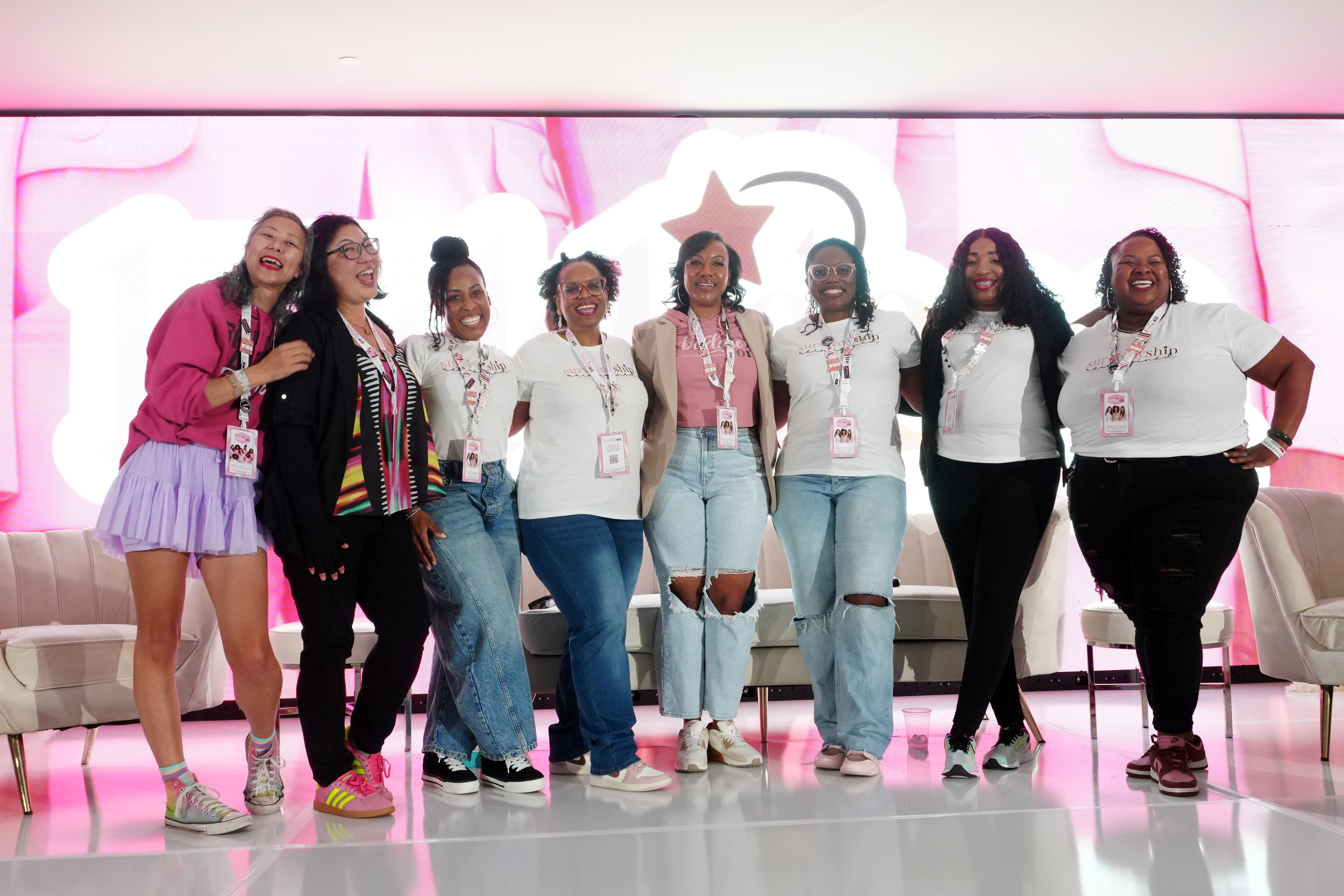 A diverse group of women standing together in front of a vibrant pink screen, smiling and engaging with each other 
